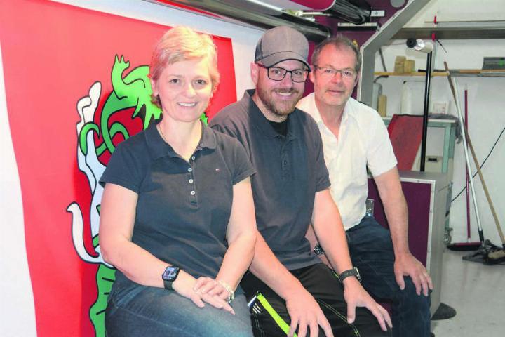 Sie sind bereits ein eingespieltes Team und arbeiten Hand in Hand: Firmengründer und jetziger Inhaber Samuel Keller und seine Nachfolger, Adrian und Monika von Arx (von rechts) vor dem Herzstück des Betriebs, dem grossen Textildrucker. Hier wird gerade eine Fahne der ehemaligen Bündner Gemeinde Schlans ausgedruckt. Bild: Chregi Hansen